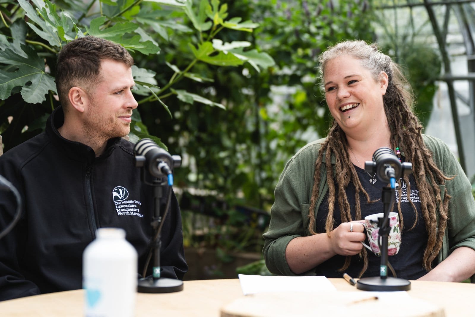 Two Lancashire Wildlife Trust staff recording an episode of Conservation Chronicles at a table among glasshouse plants.